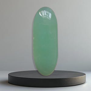 Op de afbeelding: Een lang, ovaalvormig, doorschijnend, lichtgroen object. Het object wordt getoond op een ronde, zwarte sokkel. Het oppervlak van het object reflecteert licht, wat wijst op een gepolijste afwerking. De achtergrond is neutraal grijs.