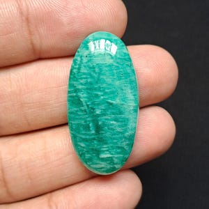 Op de afbeelding: Een ovale, gepolijste edelsteen in een opvallende teal tint, met een kenmerkend natuurlijk patroon. De steen wordt in een hand gehouden, waarbij het gladde oppervlak en de manier waarop licht met de textuur interageert, worden benadrukt.