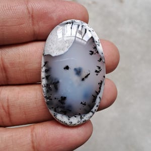 Op de afbeelding: Ovale dendritische agaat edelsteen. De steen heeft een doorschijnende witte basis met blauwe en zwarte insluitsels, die op een landschap lijken. De randen zijn een mix van wit en zwart.