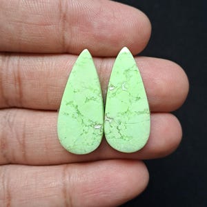 Op de afbeelding: Twee peervormige, lichtgroene edelstenen met een gemarmerd uiterlijk. De stenen hebben een glad, gepolijst oppervlak en worden tegen een donkere achtergrond gehouden. De edelstenen zijn waarschijnlijk voor het maken van sieraden.