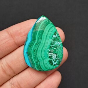 May include: A teardrop-shaped malachite gemstone with a polished surface. The stone features bands of deep green and turquoise blue, creating a swirling pattern. The gemstone is held in a hand against a dark background.