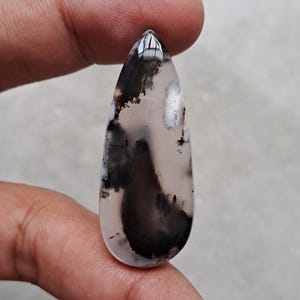 Op de afbeelding: Een druppelvormige edelsteen met een doorschijnende, bleke achtergrond en opvallende zwarte dendritische insluitsels. De gepolijste steen wordt tussen de vingers gehouden en toont zijn natuurlijke patronen. De edelsteen is ongeveer 3,8 cm lang.