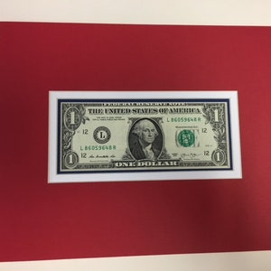 May include: A framed one-dollar bill featuring a portrait of George Washington. The bill is set against a white mat, a red border, and a cream-colored background. The bill's details are clearly visible, including the serial number and the text "ONE DOLLAR."