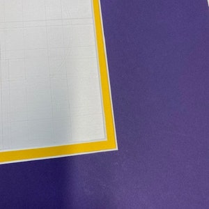 May include: A purple mat with a yellow border. The mat is on top of a white gridded sheet of paper.