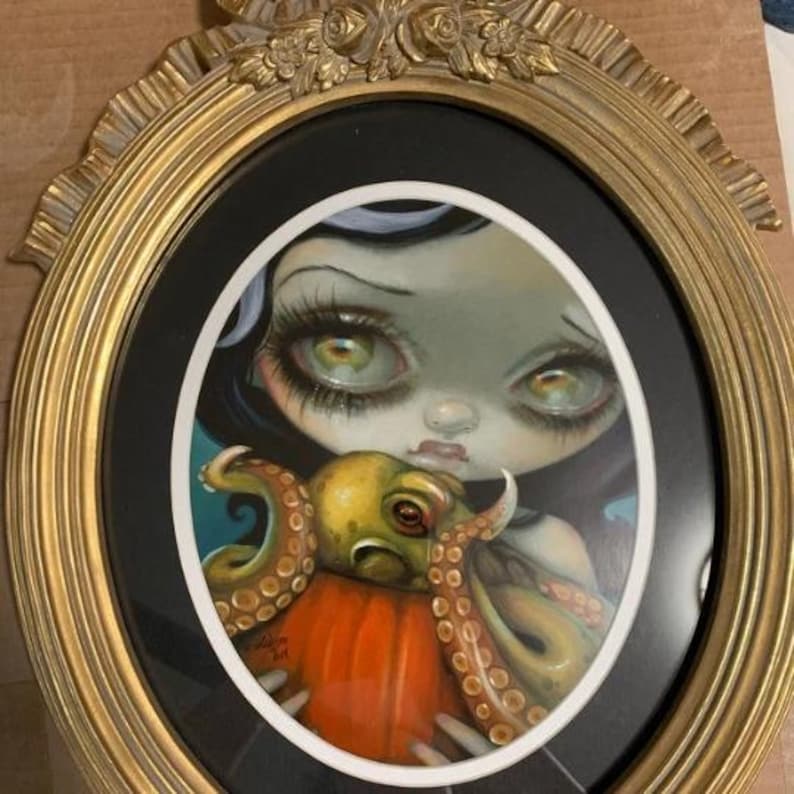K&ouml;nnte beinhalten: Oval gerahmtes Kunstwerk mit einer gro&szlig;&auml;ugigen Figur, die einen orangefarbenen K&uuml;rbis mit einem Oktopus h&auml;lt. Das Kunstwerk hat einen schwarzen Hintergrund und ist in einem goldfarbenen, verzierten Rahmen gerahmt. Die Figur hat helle Haut, schwarzes Haar und gro&szlig;e gr&uuml;ne Augen.