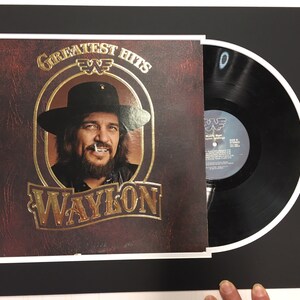 May include: A vinyl record album cover featuring a portrait of Waylon Jennings. The album cover is red and gold with the text "Greatest Hits" and "Waylon" in gold lettering. The record is black and is in a white matted frame.