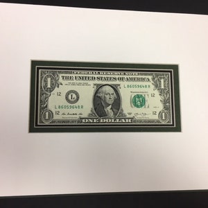 May include: A framed one dollar bill with a green border. The bill features a portrait of George Washington and the text "THE UNITED STATES OF AMERICA" and "ONE DOLLAR".