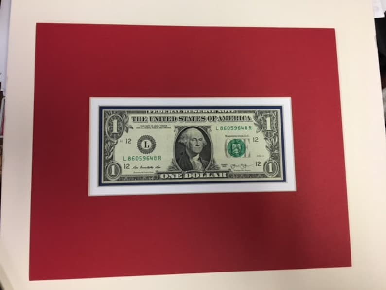 May include: A framed one dollar bill featuring a portrait of George Washington. The bill is in good condition and has a blue border. The text "THE UNITED STATES OF AMERICA" and "ONE DOLLAR" are visible.