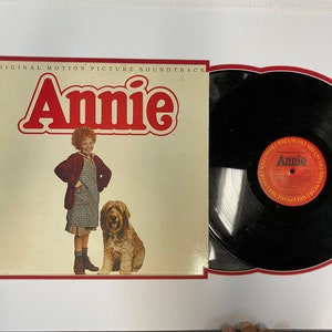 May include: A red and white vinyl record album cover with the title "Annie" in large red letters. The cover features a young girl with red hair standing next to a brown dog. The album is part of a framed set with a black vinyl record.