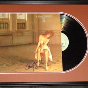 May include: A framed vinyl record album by Carly Simon, titled "No Secrets". The album cover features a woman in a white dress sitting on a chair in a room with large windows. The record is signed by Carly Simon.