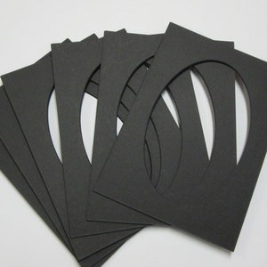 May include: A set of six rectangular black paper mats, each featuring a centered oval cutout. The mats are arranged in a fanned-out, overlapping stack, displaying their uniform dark color and precise die-cut edges for framing purposes.