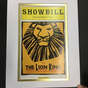 May include: A framed showbill for Disney's "The Lion King" Broadway musical. The showbill features a yellow and black image of a lion's face. The text "SHOWBILL" and "NEW AMSTERDAM THEATRE" are at the top. The text "THE LION KING" and "BROADWAY'S AWARD-WINNING BEST MUSICAL" are at the bottom.