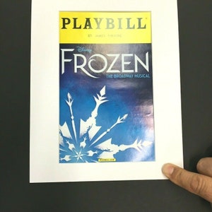 May include: A yellow and blue Playbill for the Broadway musical "Frozen" at the St. James Theatre. The Playbill features a silhouette of the two main characters, Elsa and Anna, against a blue background with a snowflake design.