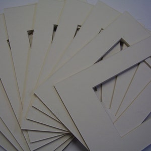 May include: A stack of ten white mat boards with a rectangular opening in the center of each board.