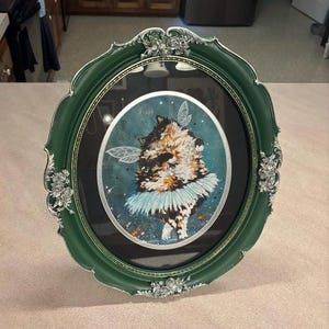 K&ouml;nnte beinhalten: Ein oval gerahmtes Kunstwerk mit einer Katze mit Feenfl&uuml;geln und einem blauen Tutu. Der Rahmen ist dunkelgr&uuml;n mit silbernen Akzenten. Die Katze hat braunes, schwarzes und wei&szlig;es Fell, vor einem t&uuml;rkisfarbenen Hintergrund.