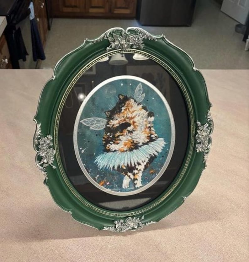 K&ouml;nnte beinhalten: Ein oval gerahmtes Kunstwerk mit einer Katze mit Feenfl&uuml;geln und einem blauen Tutu. Der Rahmen ist dunkelgr&uuml;n mit silbernen Akzenten. Die Katze hat braunes, schwarzes und wei&szlig;es Fell, vor einem t&uuml;rkisfarbenen Hintergrund.