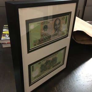May include: Two framed Vietnamese banknotes, each featuring a portrait of a man and the text "Cộng H&ograve;a X&atilde; Hội Chủ Nghĩa Việt Nam" and "MỘT TR&Aacute;M NGH&Igrave;N ĐỒNG" with the denomination "100.000" printed in green.