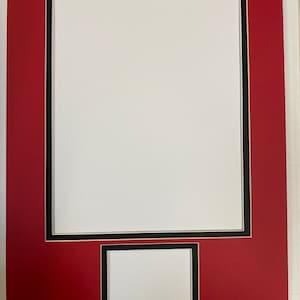 May include: A red picture mat with a black and white border, featuring two openings for photos or artwork. The larger opening is rectangular, and the smaller one is below it. The mat is designed for framing.