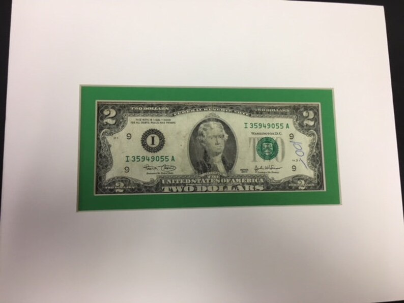 May include: A framed two dollar bill with a green border. The bill features a portrait of Thomas Jefferson and the text "I 35949055 A" printed on it.