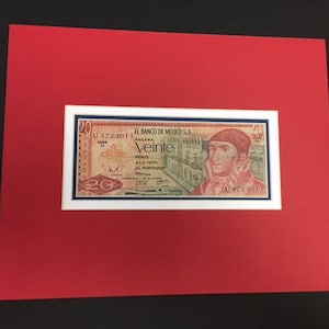 May include: A framed 20 peso bill from the Banco de Mexico, S.A. The bill features a portrait of a man in a military uniform, with the text "Veinte" in red and the words "Banco de Mexico, S.A." in blue.