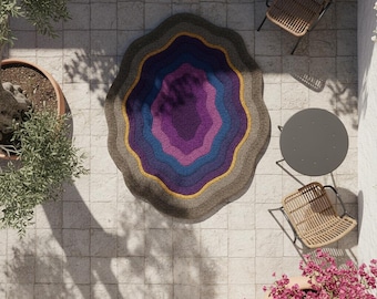 Puede incluir: Una alfombra redonda con un diseño en capas en tonos morados, azules y grises, con un acento amarillo. La alfombra tiene un borde ondulado y está colocada en un patio embaldosado. El diseño de la alfombra se asemeja a una sección transversal de una geoda.