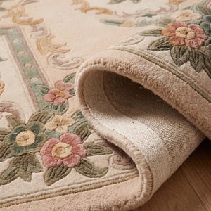 Puede incluir: Alfombra beige con diseño floral, parcialmente enrollada, revelando el reverso. El diseño presenta un borde floral repetitivo y un patrón floral central en rosa, verde y amarillo. La alfombra está hecha de un material suave y texturizado.