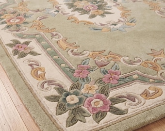 Puede incluir: Una alfombra rectangular con un fondo verde claro y un diseño floral. La alfombra presenta un borde y un panel central de color crema, con patrones florales rosas, amarillos y verdes. La alfombra está colocada sobre un suelo de madera.
