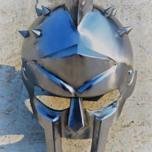 May include: A silver-colored gladiator helmet with a prominent crest and several pointed spikes. The helmet features a face guard with eye and mouth openings and is made of a reflective metal.