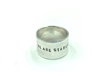 May include: A wide, silver ring with the words "WE ARE STARDUST" engraved around the band. The ring has a simple, minimalist design and is set against a white background.