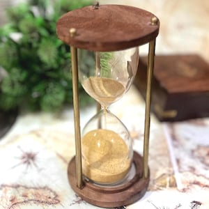 May include: A vintage hourglass with a dark brown wooden frame and base, and clear glass. The hourglass contains tan sand. The hourglass is set on a map with a compass rose design. The hourglass is a decorative item.