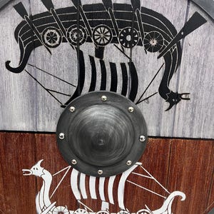 Puede incluir: Un escudo circular de madera con un umbo central de metal oscuro fijado con remaches. La mitad superior es de color gris claro con un gr&aacute;fico de un barco vikingo negro, mientras que la mitad inferior es marr&oacute;n oscuro con un gr&aacute;fico blanco.