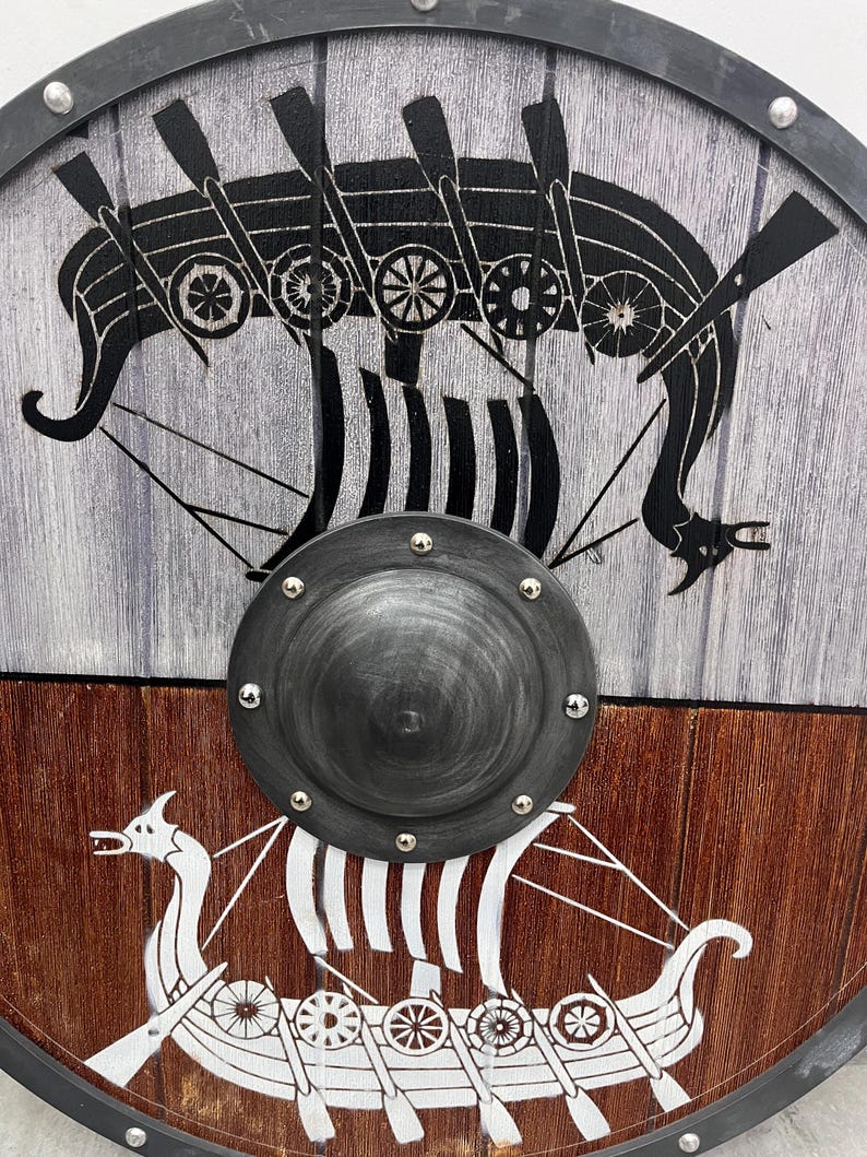 Puede incluir: Un escudo circular de madera con un umbo central de metal oscuro fijado con remaches. La mitad superior es de color gris claro con un gr&aacute;fico de un barco vikingo negro, mientras que la mitad inferior es marr&oacute;n oscuro con un gr&aacute;fico blanco.
