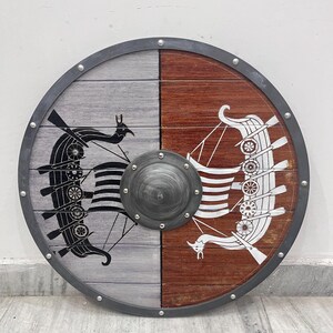 Puede incluir: Un escudo circular de madera con un dise&ntilde;o dividido, con el lado izquierdo gris y el derecho marr&oacute;n. Un barco vikingo negro est&aacute; pintado en el lado gris, mientras que un barco vikingo blanco est&aacute; en el lado marr&oacute;n. Un umbo met&aacute;lico se sit&uacute;a en el centro.