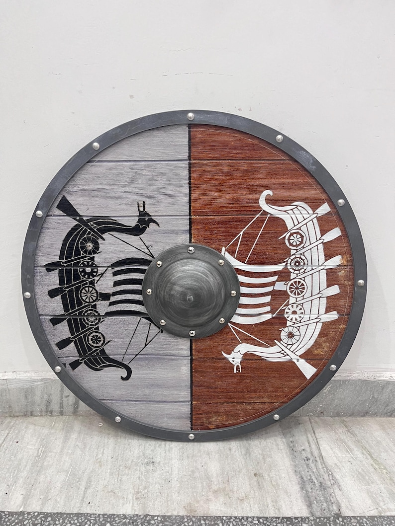 Puede incluir: Un escudo circular de madera con un dise&ntilde;o dividido, con el lado izquierdo gris y el derecho marr&oacute;n. Un barco vikingo negro est&aacute; pintado en el lado gris, mientras que un barco vikingo blanco est&aacute; en el lado marr&oacute;n. Un umbo met&aacute;lico se sit&uacute;a en el centro.