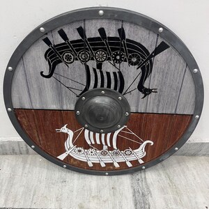 Puede incluir: Un escudo circular de madera con un borde met&aacute;lico con remaches y un umbo central de metal. La mitad superior es de color gris claro con un gr&aacute;fico de barco vikingo negro, mientras que la mitad inferior es marr&oacute;n oscuro con un gr&aacute;fico de barco blanco.