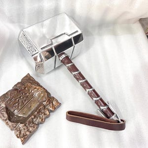 Könnte beinhalten: Ein silberner Hammer mit braunem Ledergriff und Riemen. Der Hammerkopf hat dekorative Gravuren. Ein brauner, quadratischer Ständer ist ebenfalls vorhanden. Replik.