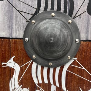 Puede incluir: Un escudo circular de madera con un umbo central de metal gris oscuro fijado con seis remaches plateados. La superficie del escudo est&aacute; dividida en paneles horizontales y muestra un gr&aacute;fico de un barco vikingo pintado en blanco sobre madera.