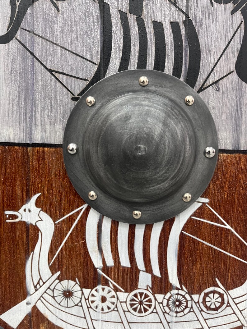 Puede incluir: Un escudo circular de madera con un umbo central de metal gris oscuro fijado con seis remaches plateados. La superficie del escudo est&aacute; dividida en paneles horizontales y muestra un gr&aacute;fico de un barco vikingo pintado en blanco sobre madera.