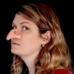 May include: A prosthetic witch nose made of flesh-toned material with a small, round nostril. The nose is worn on a person's face, with the person's hair and ear visible. The background is black.