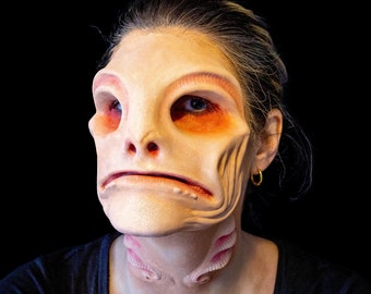 May include: A prosthetic mask in a pale flesh tone, featuring exaggerated facial features and a textured surface. The mask includes large eye sockets, a prominent nose, and a downturned mouth. The neck area has a textured design. The background is black.