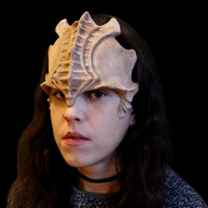 May include: A beige, textured mask with a detailed, ridged design covering the forehead and eye area. The mask has a weathered appearance, suggesting a vintage or fantasy theme. The mask is worn by a person.