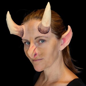May include: A prosthetic devil horn and ear set. The horns are a light tan color with a darker brown base. The ears are pink and flesh-toned. The nose is also flesh-toned. The set is designed to be worn on the face.