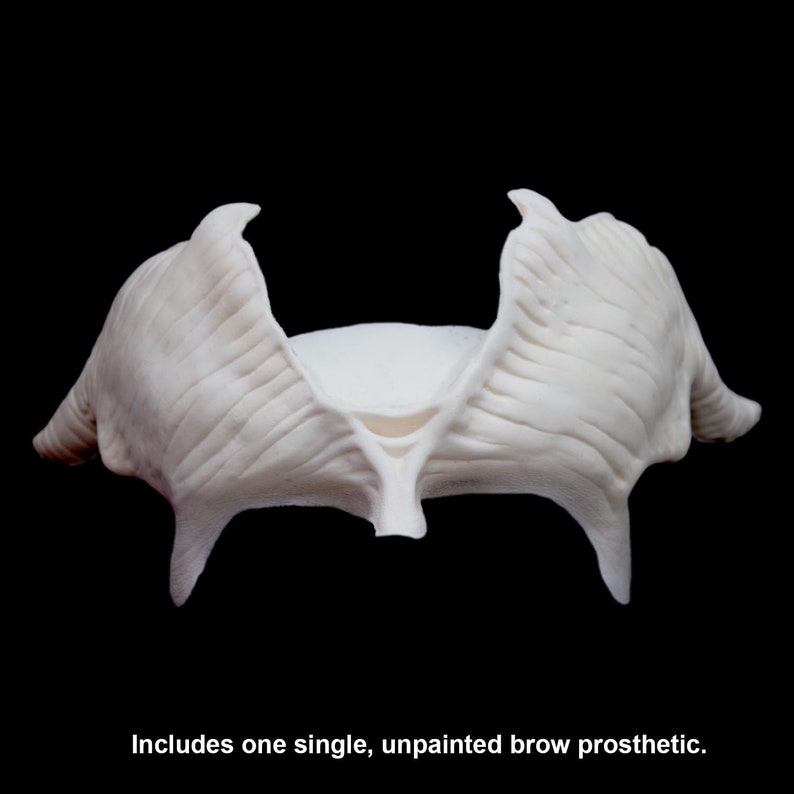 May include: A single, unpainted brow prosthetic made of a light-colored material. The prosthetic has a textured surface and is designed to be applied to the forehead. The image is set against a black background.
