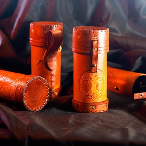 May include: Four cylindrical tan leather cases with lids, featuring embossed tree of life designs. Each case has stitched edges, metal rivets, and a vertical strap closure. The leather has a smooth, polished finish with visible grain and natural color variations.