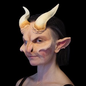 May include: A prosthetic mask featuring a demonic face with large, curved horns, pointed ears, and a prominent nose. The mask is painted in shades of beige, pink, and brown, with a black background.
