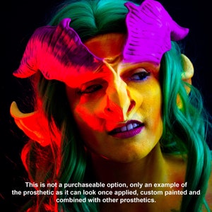 May include: A person with a prosthetic face and horns, painted with bright colors. The face is yellow and orange, with purple horns and green hair. The image is an example of how the prosthetic can look when applied and painted.