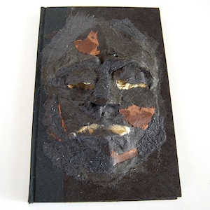 May include: A black book cover with a textured, three-dimensional face made of paper and fabric. The face has a distressed look with brown and gold accents.