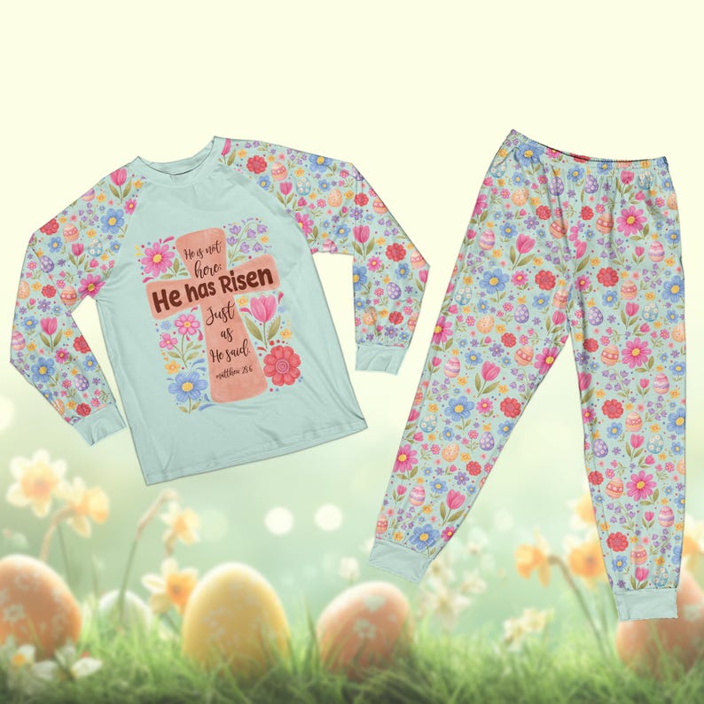 May include: A light blue Easter-themed pajama set. The long-sleeved top features a cross design with the text "He has Risen". The sleeves and pajama pants are covered in a floral and Easter egg pattern. The pajama set is made of a soft, comfortable material.