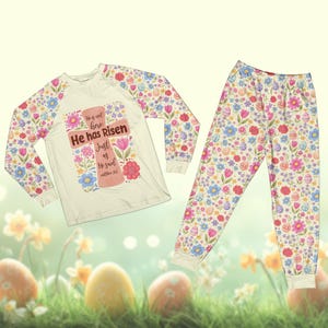 May include: Easter-themed pajama set. The long-sleeved top features a cross design with the text "He has Risen". The sleeves and pajama pants are covered in a floral and Easter egg pattern. The set is cream-colored with pastel accents.