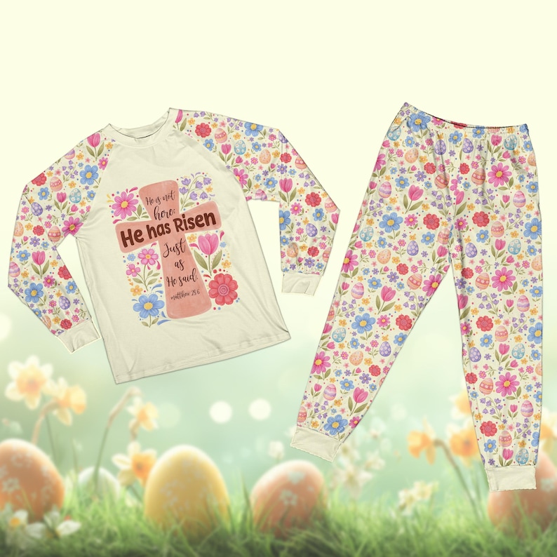 May include: Easter-themed pajama set. The long-sleeved top features a cross design with the text "He has Risen". The sleeves and pajama pants are covered in a floral and Easter egg pattern. The set is cream-colored with pastel accents.
